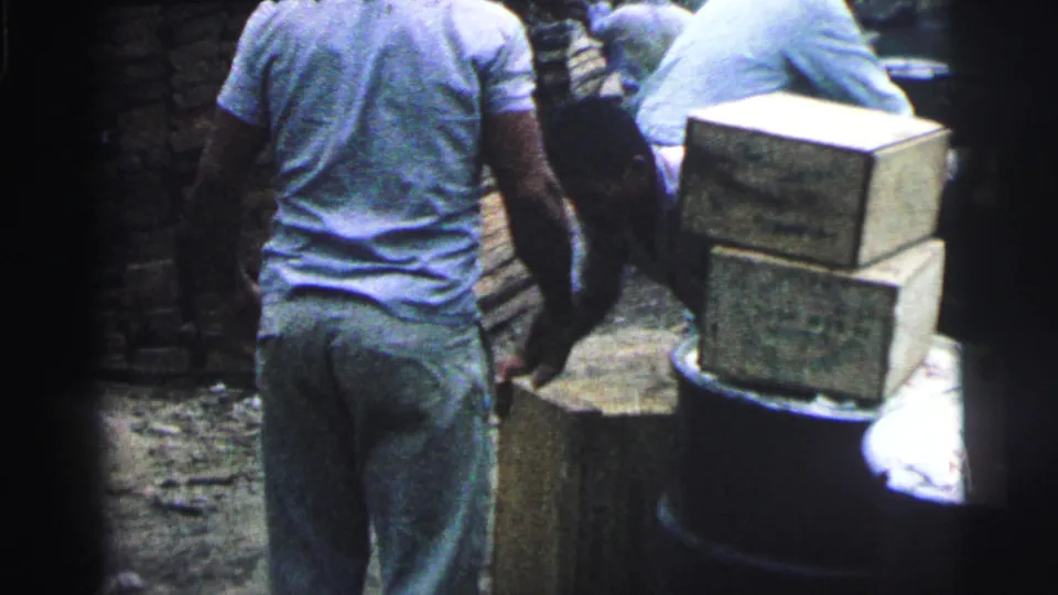 Handheld 8mm view of a worker from behind beside wooden crates and barrels in a Brazilian dock setting.