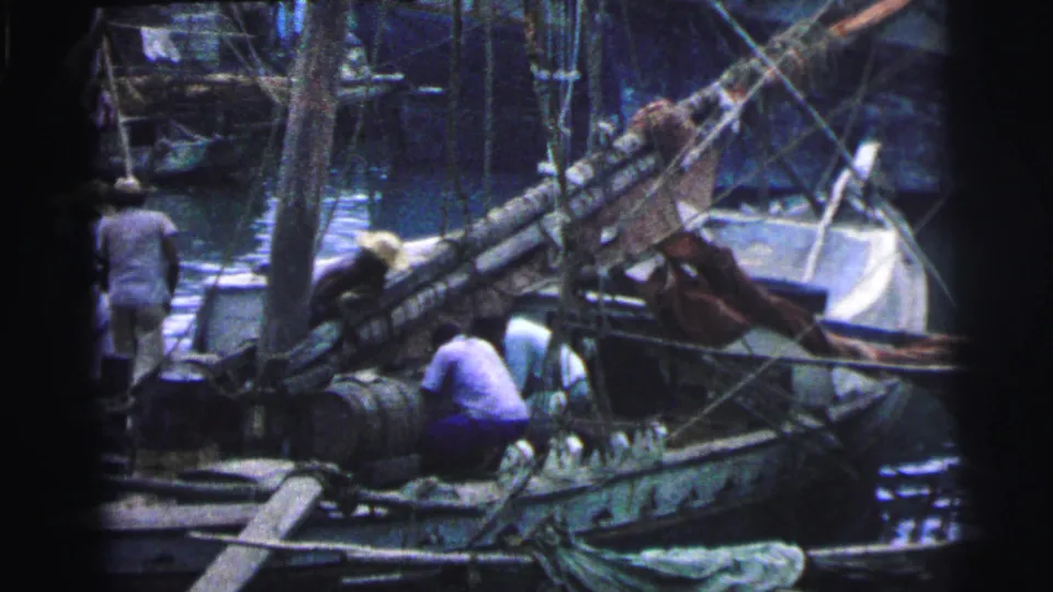 Wider 1967 Brazil port frame with workers near barrels, ropes, and a traditional sailboat at the water's edge.