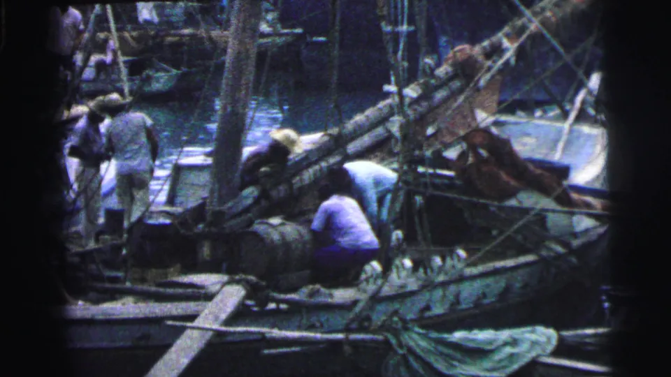 1967 Brazil dockside frame with men preparing a small fishing boat among rigging, barrels, planks, and reflective harbor water.