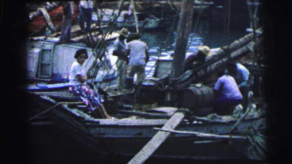1967 Brazil waterfront frame with a seated woman, workers, wooden boats, rigging, barrels, and harbor water in view.
