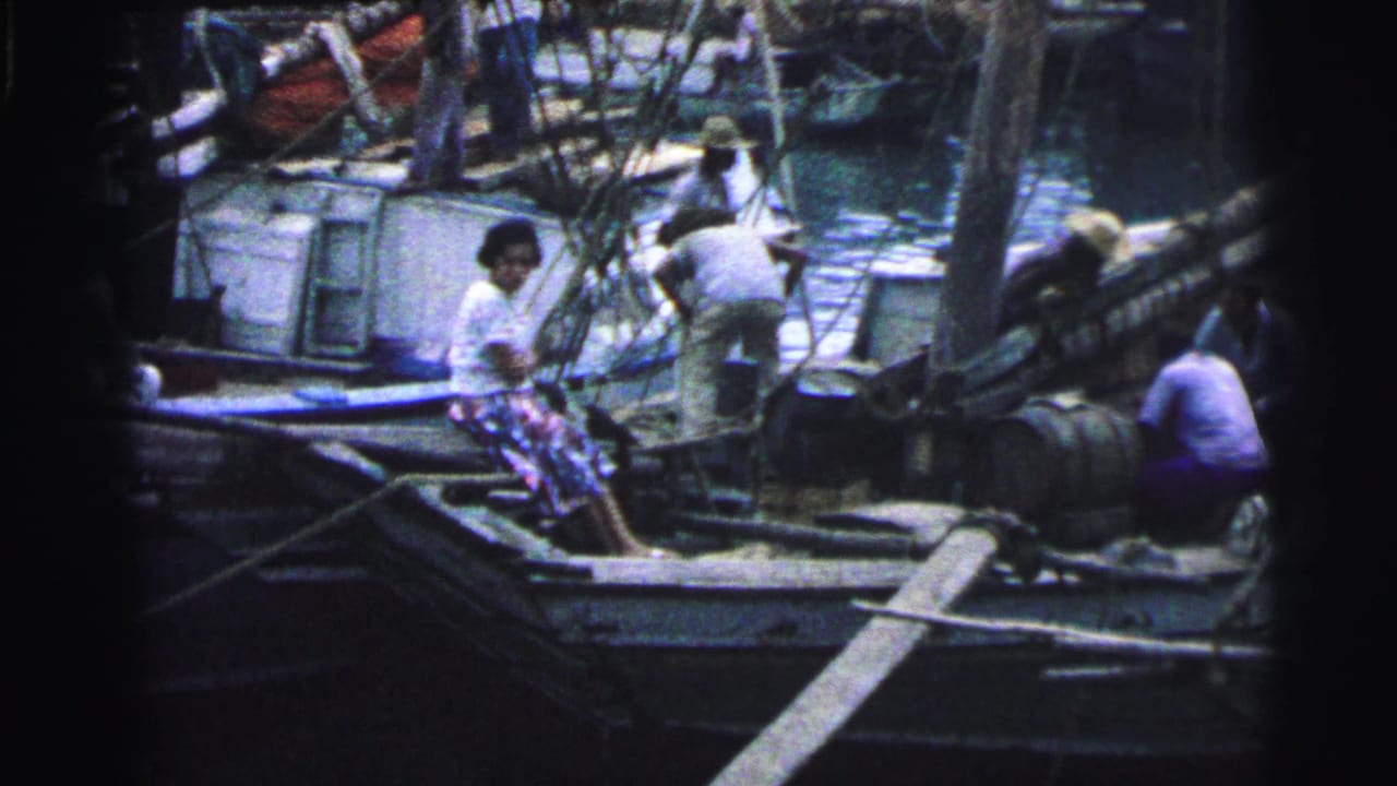 People sit and work on wooden boats beside water, with ropes and barrels nearby.