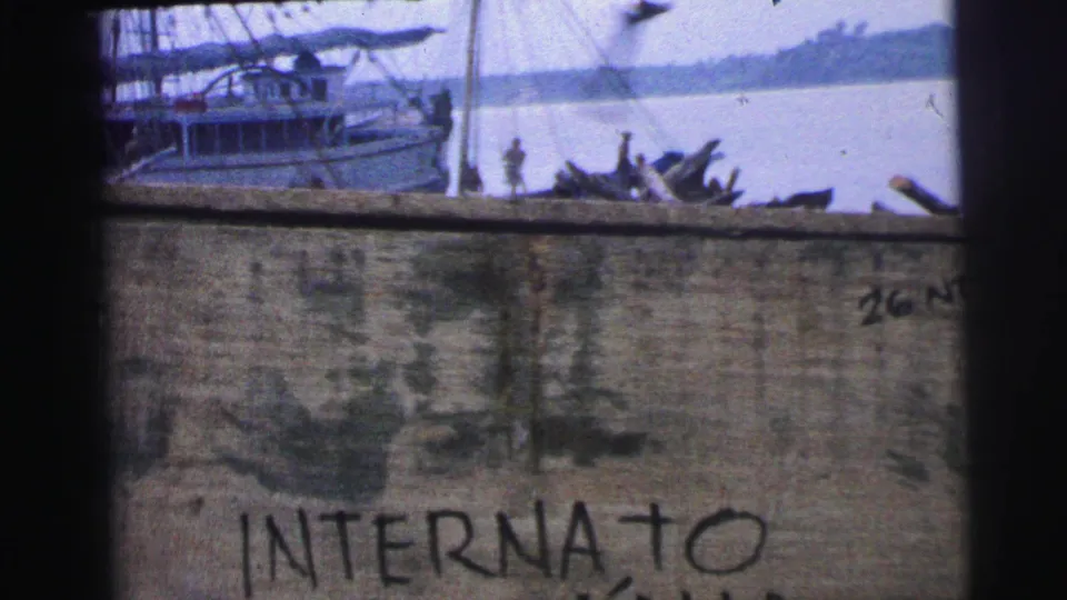 1967 Brazil 8mm frame with a fishing boat on calm water behind a weathered wall carrying partial handwritten text.