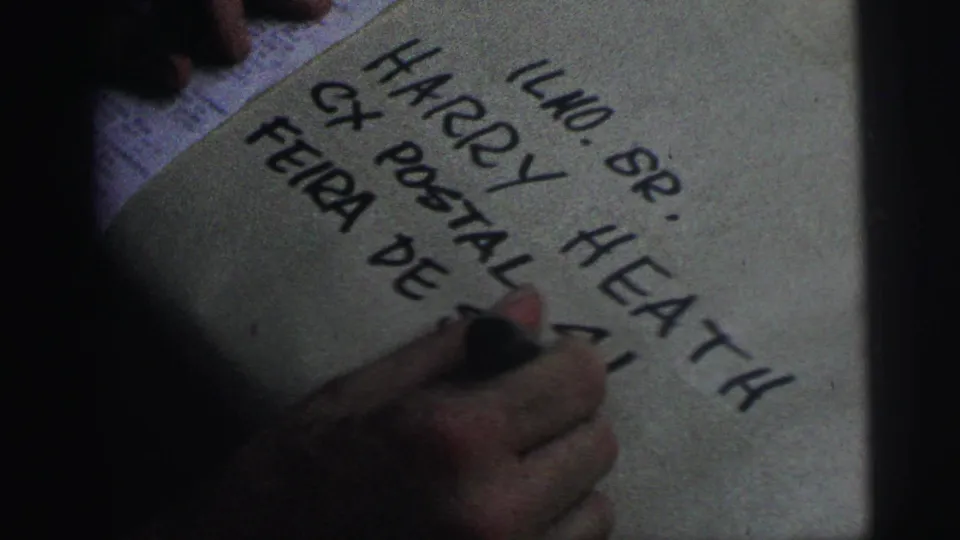 1967 Brazil 8mm frame showing a hand writing large block letters on a pale surface, with “HARRY HEATH” and “CX POSTAL” visible.