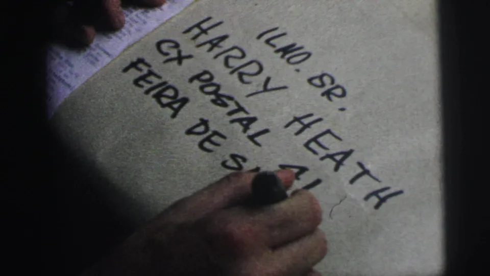1967 Brazil 8mm frame showing a hand writing an address on a paper-wrapped package, with Harry Heath and postal text partly visible.