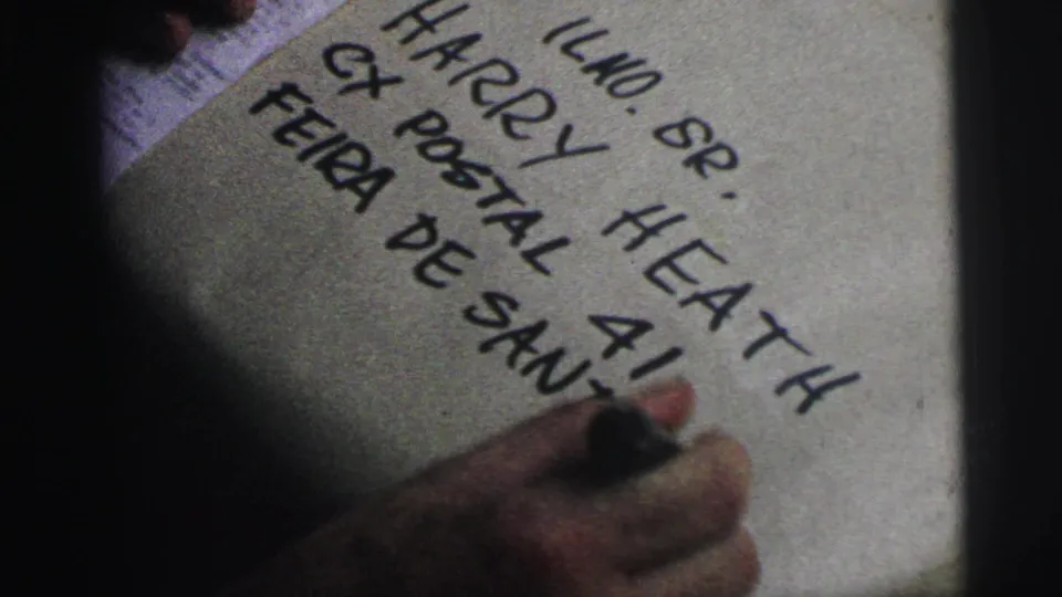 Close 8mm view of a hand adding letters to a package address, including visible Harry Heath and postal box wording.