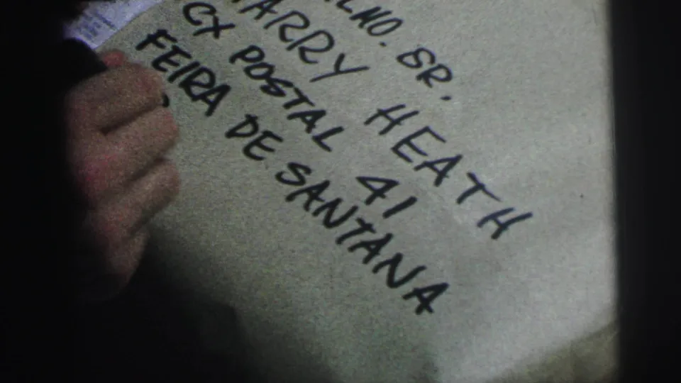 1967 Brazil home-movie frame with a hand and marker beside handwritten address text ending in Feira de Santana.