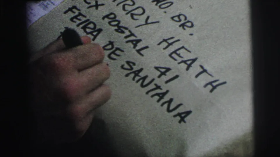 Close 8mm frame from Brazil showing a hand over a completed package address with Harry Heath, postal box, and Feira de Santana visible.