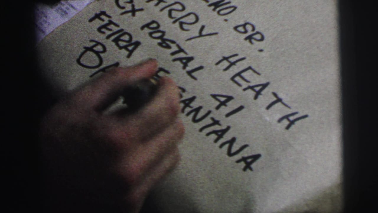 Close 1967 Brazil home-movie frame of a hand writing address text on a paper-wrapped package with a black marker.