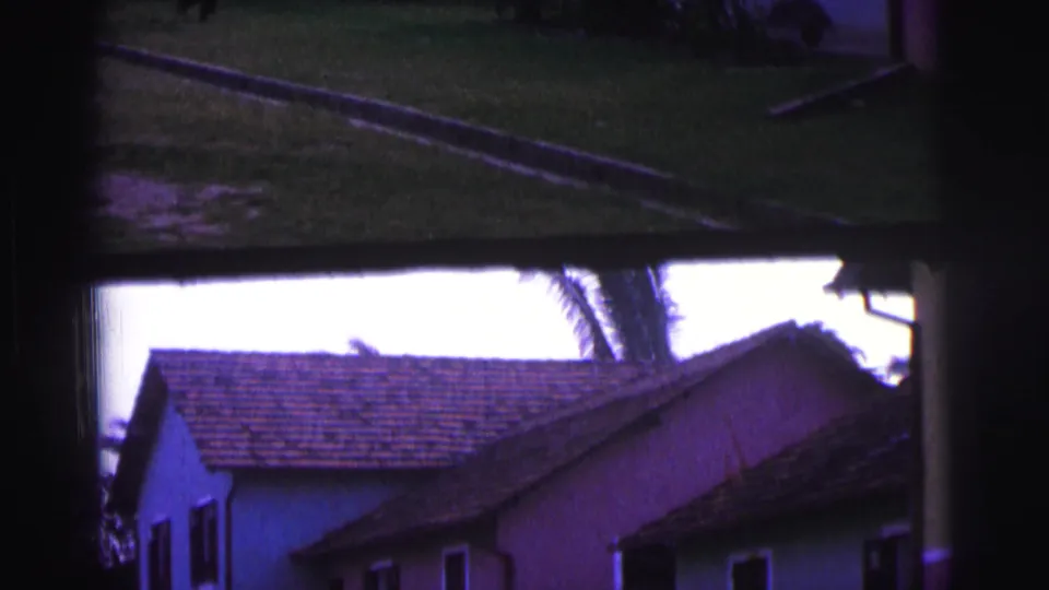 1967 Brazil 8mm frame showing pastel residential buildings with tiled roofs and palm fronds, partly obscured by dark framing.