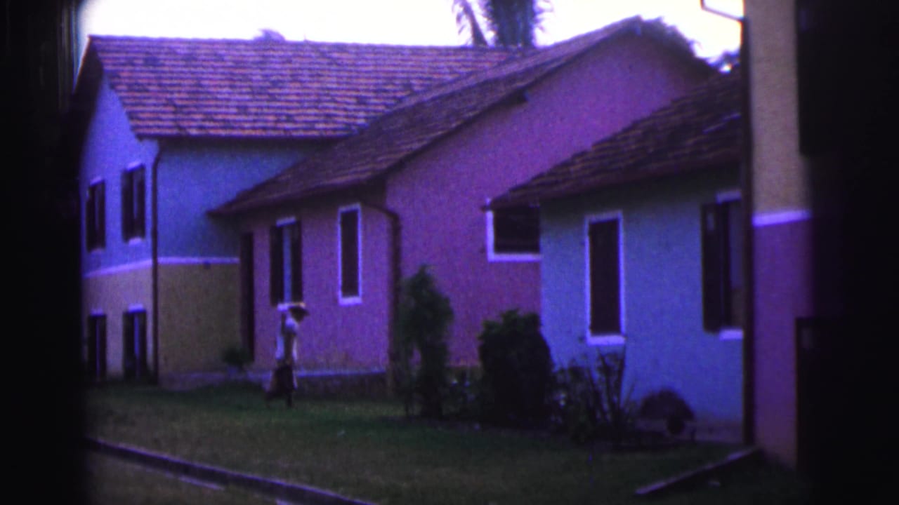 1967 Brazil 8mm frame showing colorful tiled-roof homes, lawn, palms, and a person walking near the buildings.