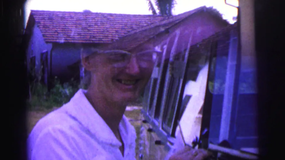 1967 Brazil 8mm frame of a smiling woman in glasses beside a vehicle, with pastel homes and foliage in the background.