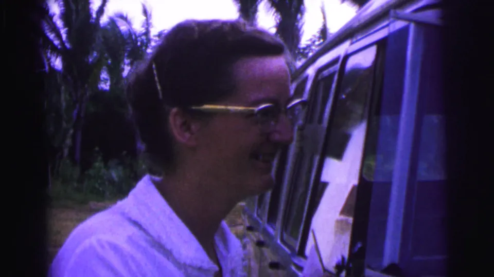 1967 Brazil 8mm frame showing a smiling man in glasses next to a vehicle, with palm trees and residential foliage behind.
