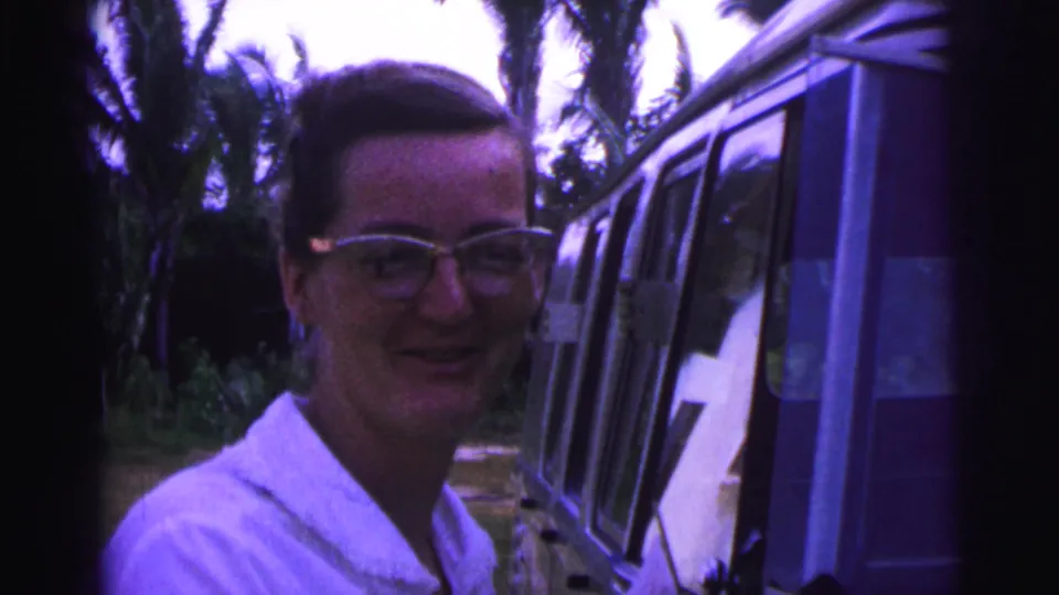 A woman in glasses turns toward the camera beside a van, with palm-like trees behind her in 1967 Brazil footage.