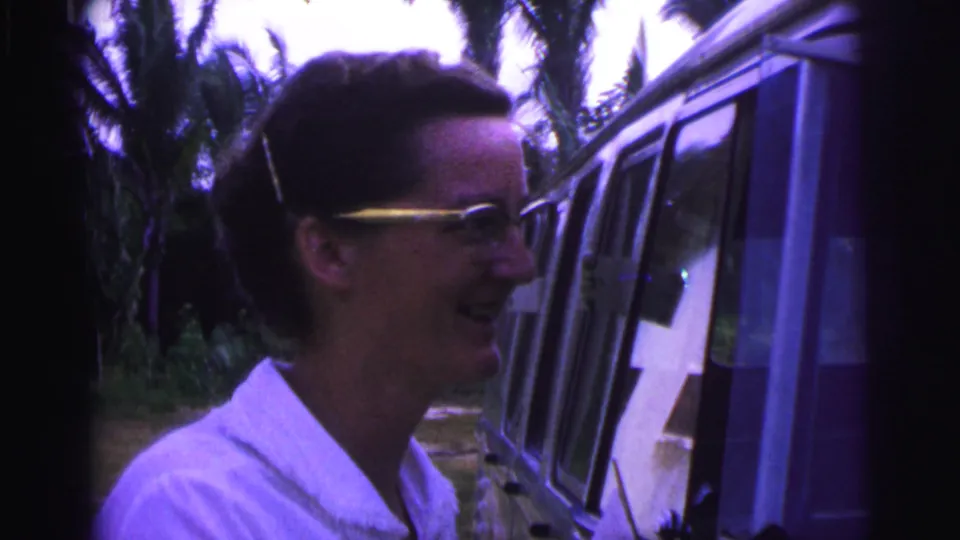 The woman smiles in profile beside a vintage van, framed by tropical greenery in archival 8mm footage from Brazil.
