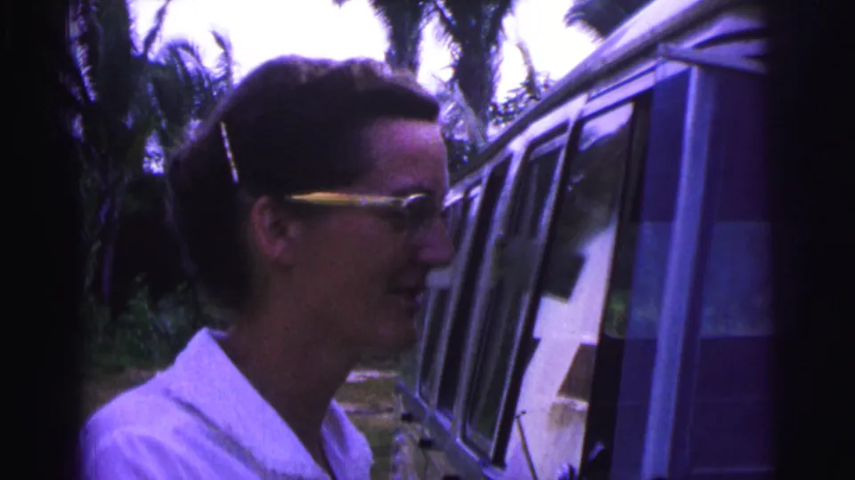 The woman appears to speak while standing in profile near the van, with trees and shaded foliage behind her in the 1967 Brazil clip.