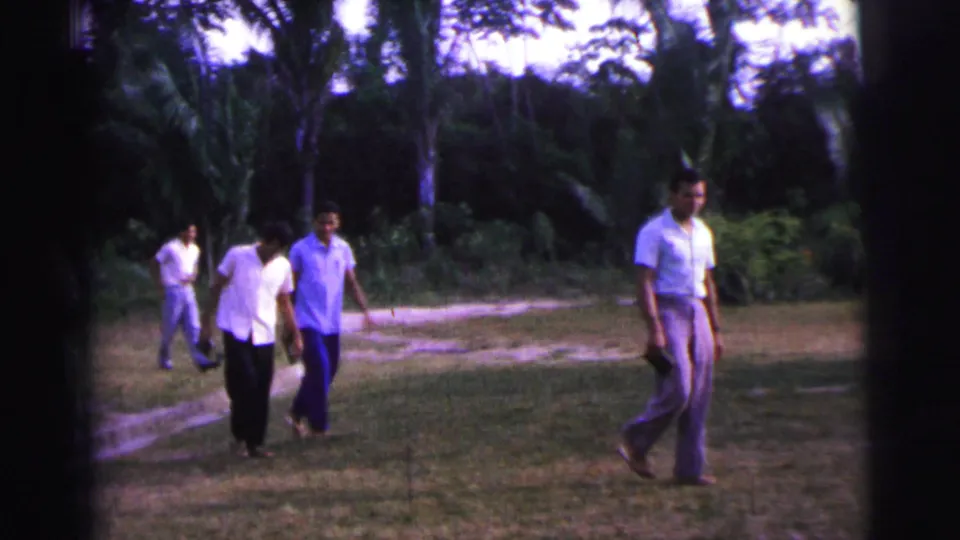 In 1967 Brazil home-movie footage, several adult men cross a lawn in a tropical garden setting with trees and pale footpaths.