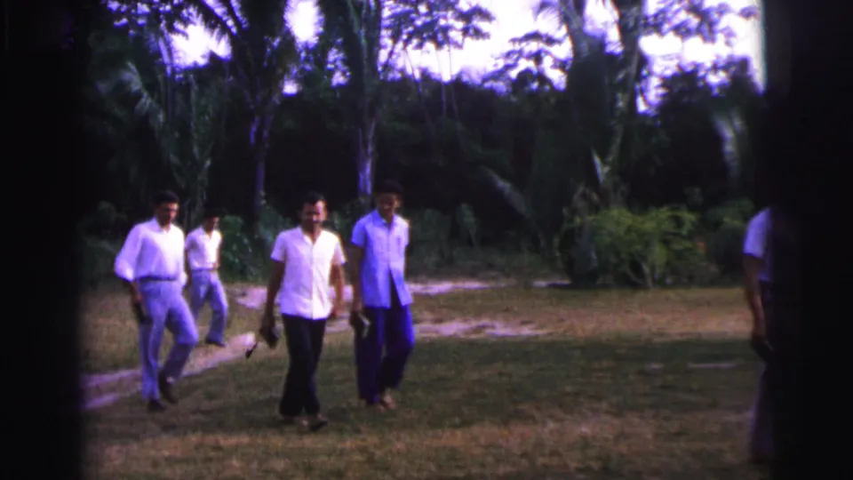 Adult men walk toward the foreground across a grassy area in 1967 Brazil, with palm-like trees and dense greenery behind them.