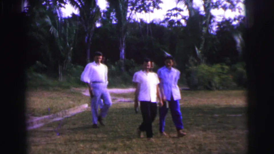 Three adult men in casual shirts and trousers pass near the camera in Brazil, with another man farther back on the lawn.