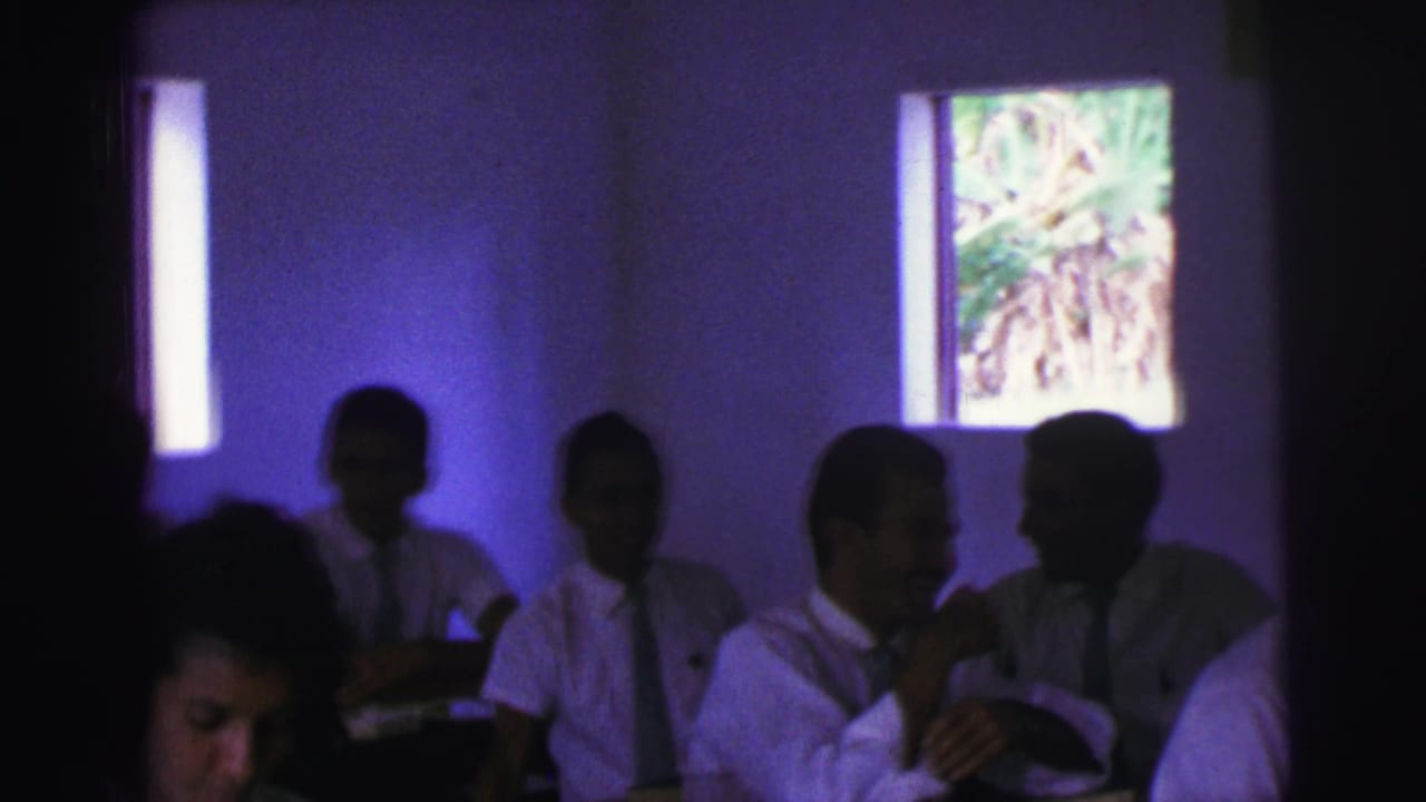 Teenagers in white shirts and ties sit at classroom desks near narrow windows.