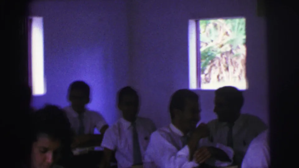 In a 1967 Brazil classroom scene, uniformed teenagers sit at wooden desks while bright windows reveal foliage outside.