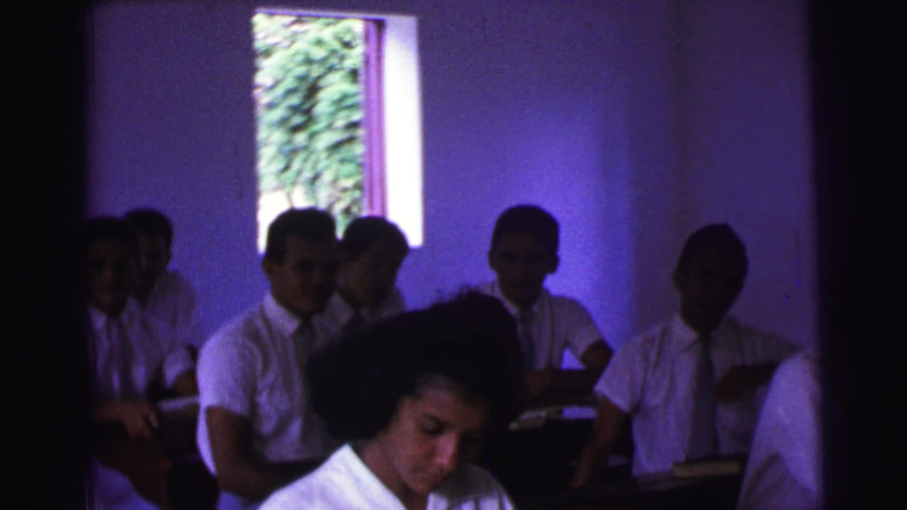A dim 1967 Brazil classroom view shows students in white shirts and ties seated at wooden desks as one foreground student looks down.