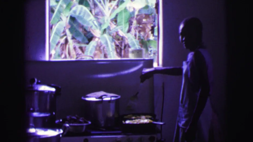 The woman appears in profile in a dim Brazilian kitchen, reaching toward a pan on the stove while pots sit below a window filled with banana leaves.