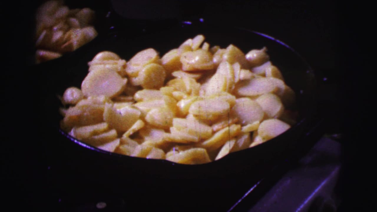 Close-up from the 8mm home movie shows sliced potatoes cooking in a dark pan, with the surrounding stovetop mostly in shadow.