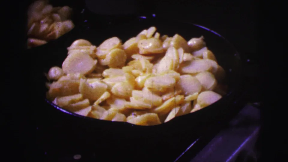 Close-up from the 8mm home movie shows sliced potatoes cooking in a dark pan, with the surrounding stovetop mostly in shadow.