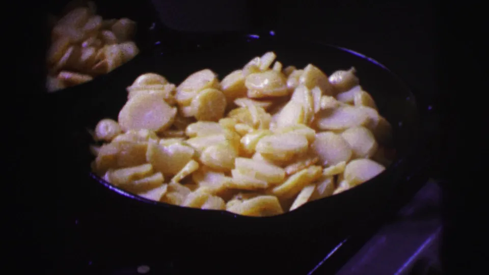Close 8mm view of pale sliced potatoes in a dark pan, with the stove and room mostly in shadow.