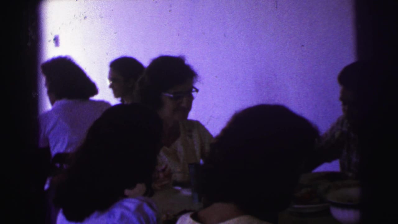 1967 Brazil home-movie frame of adults gathered around a table, silhouetted against a purple-tinted interior wall.