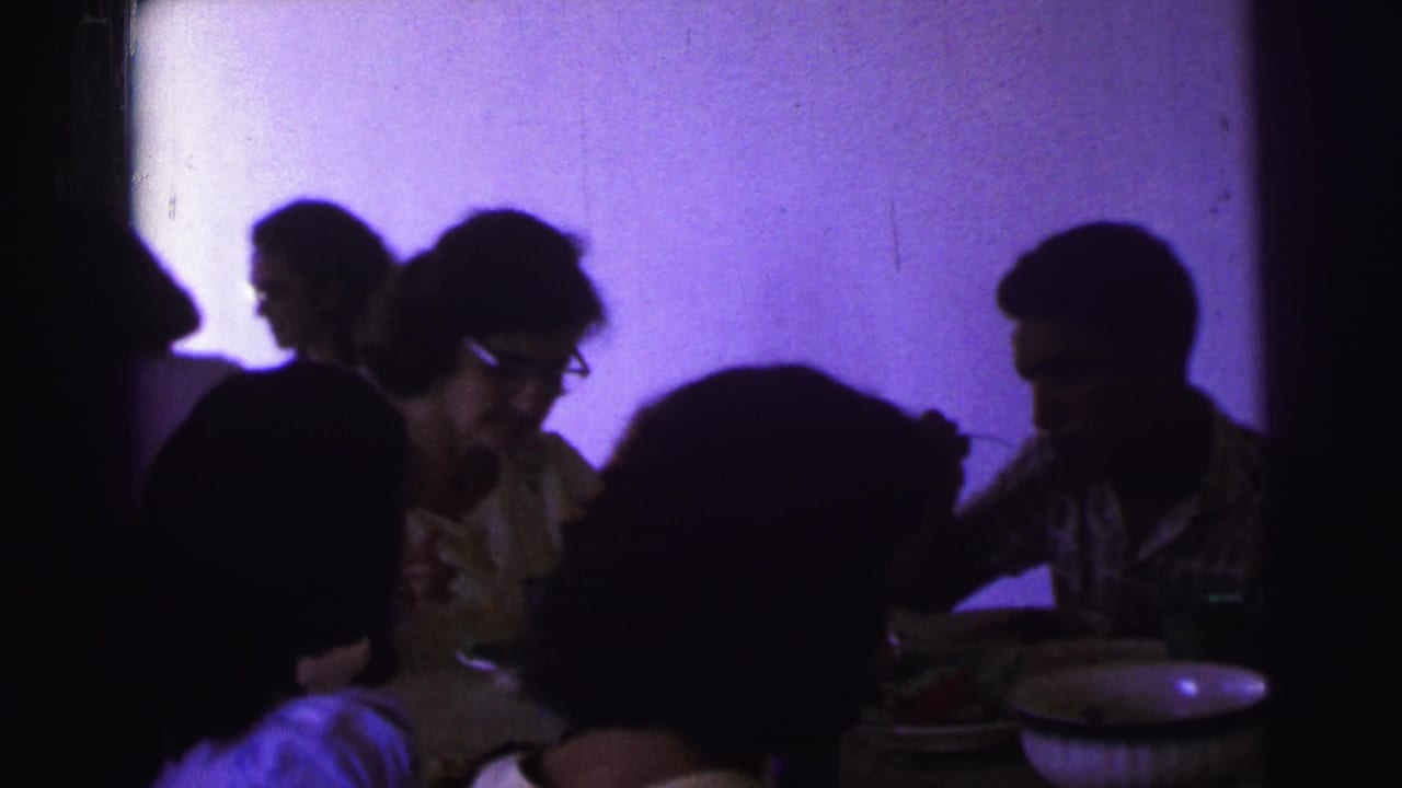 1967 Brazil home-movie frame showing adults gathered at a dinner table under strong purple indoor light.