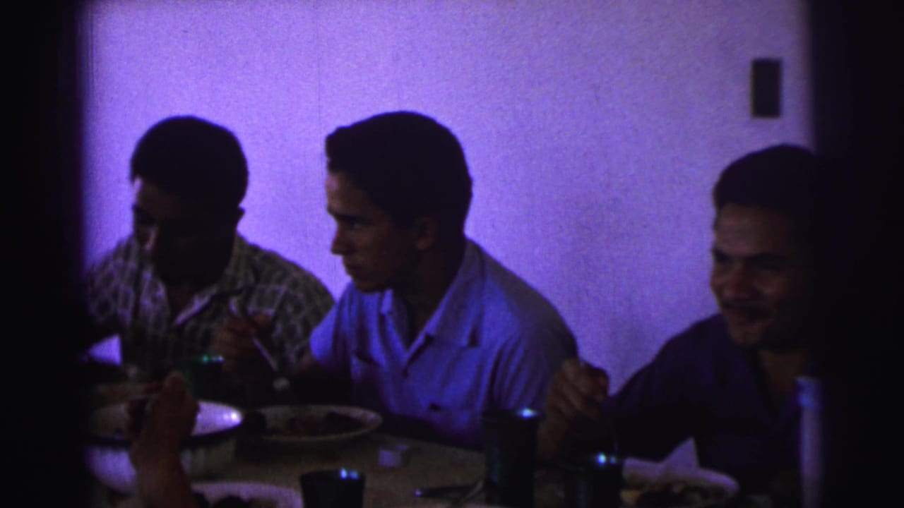 Three men sit at a dining table with plates, glasses, and utensils in a dim room.