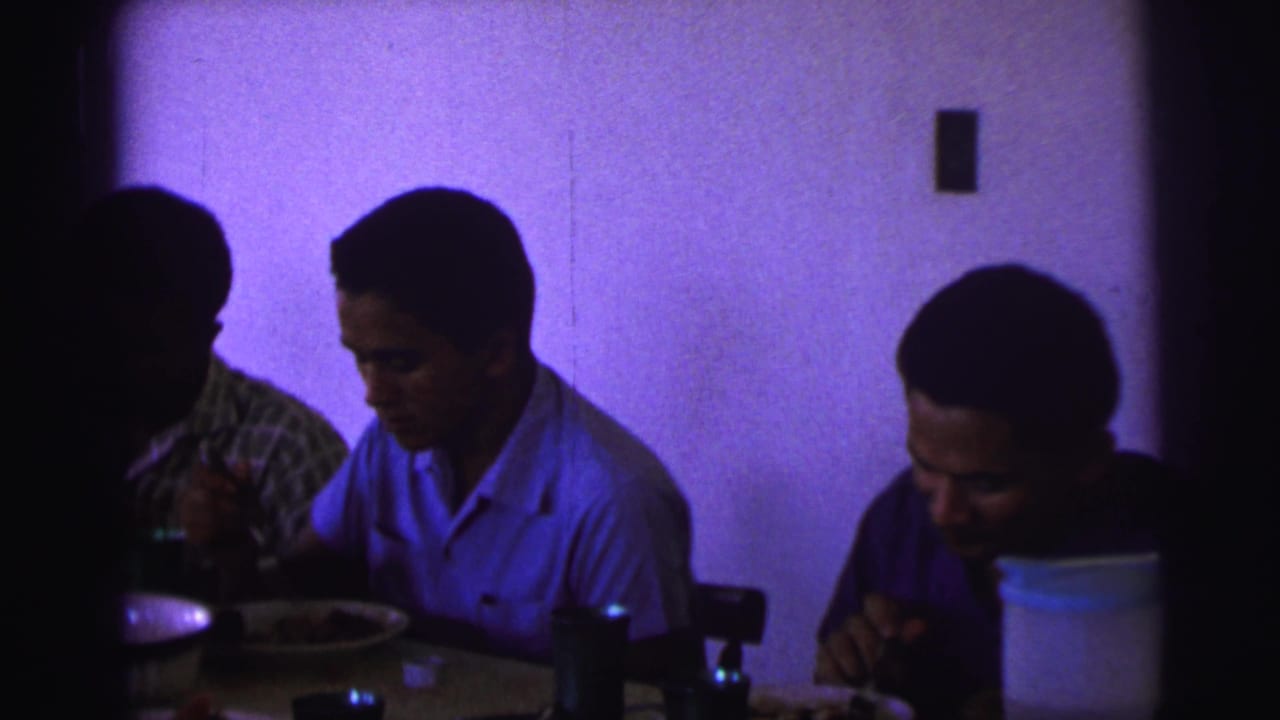Three men in casual shirts eat at an indoor table, with a pitcher and plates visible in the 1967 Brazil home-movie frame.
