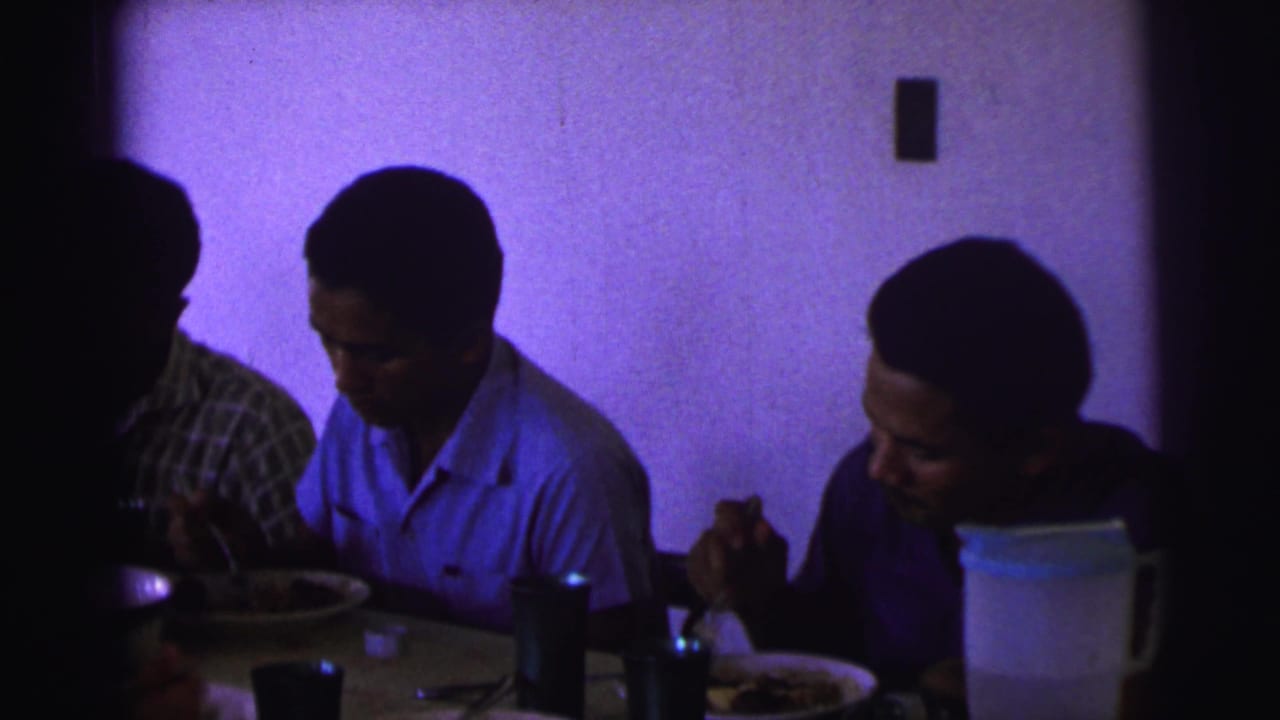 A dim 1967 Brazil dining scene shows two men eating at the table while another person is partly obscured by shadow.