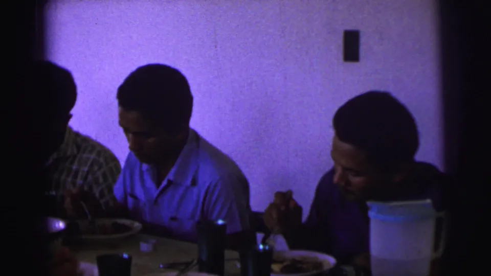 A dim 1967 Brazil dining scene shows two men eating at the table while another person is partly obscured by shadow.
