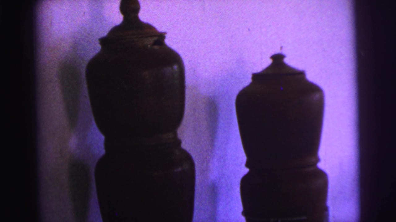 Two dark lidded ceramic pots appear against a plain interior wall, shifting the 1967 Brazil home movie from diners to household objects.