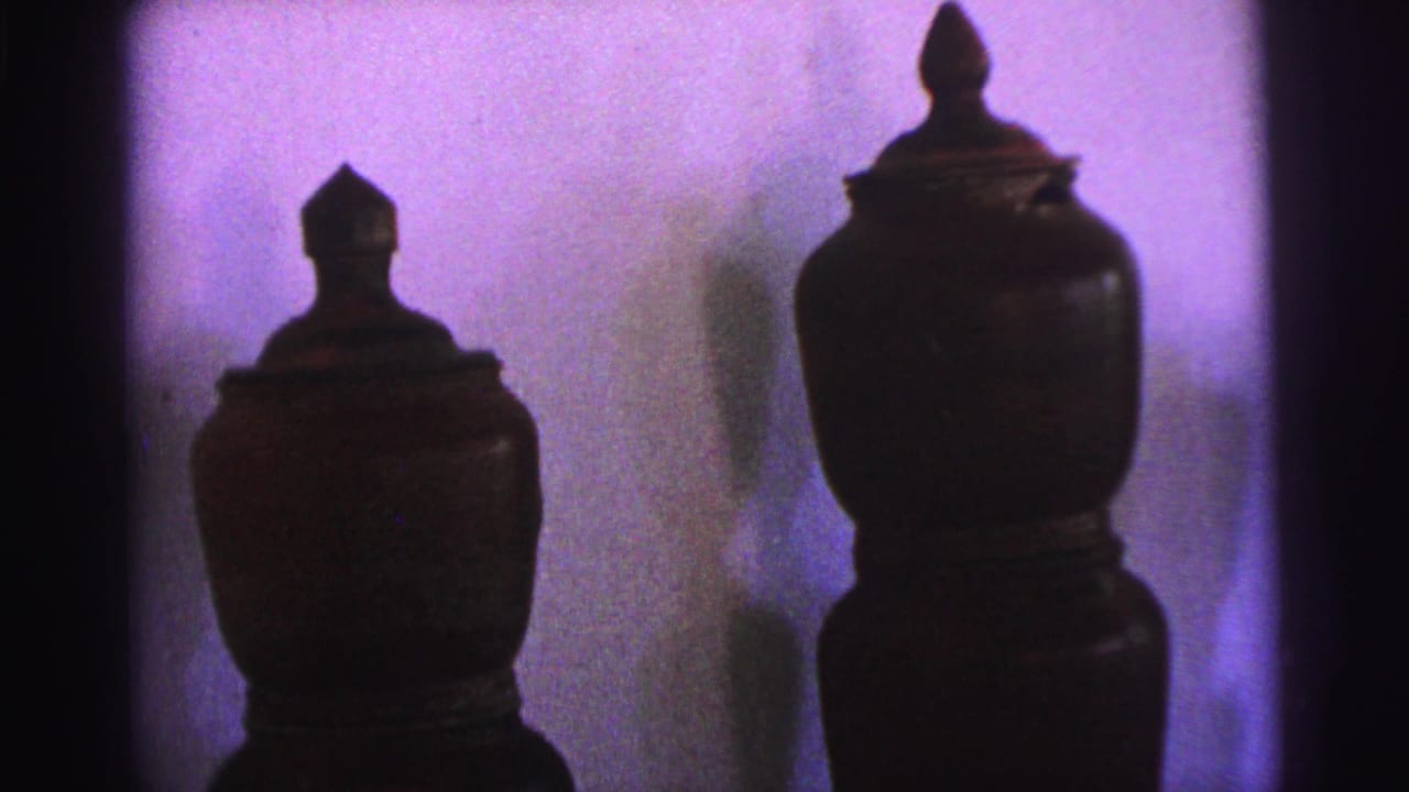 1967 Brazil home-movie frame showing two lidded brown ceramic pots on stands, with strong shadows and a purple wall cast.
