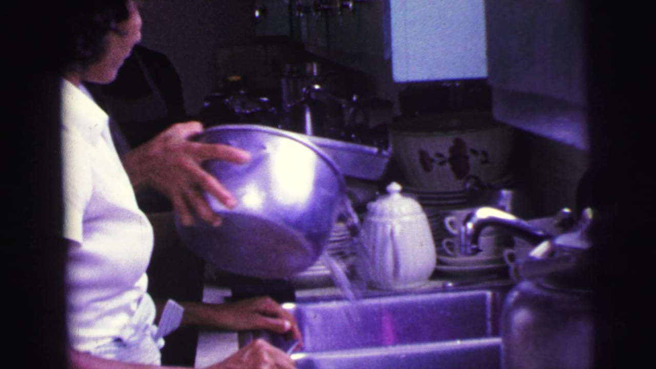 Woman in a white shirt pours water from a metal bowl into a kitchen sink.