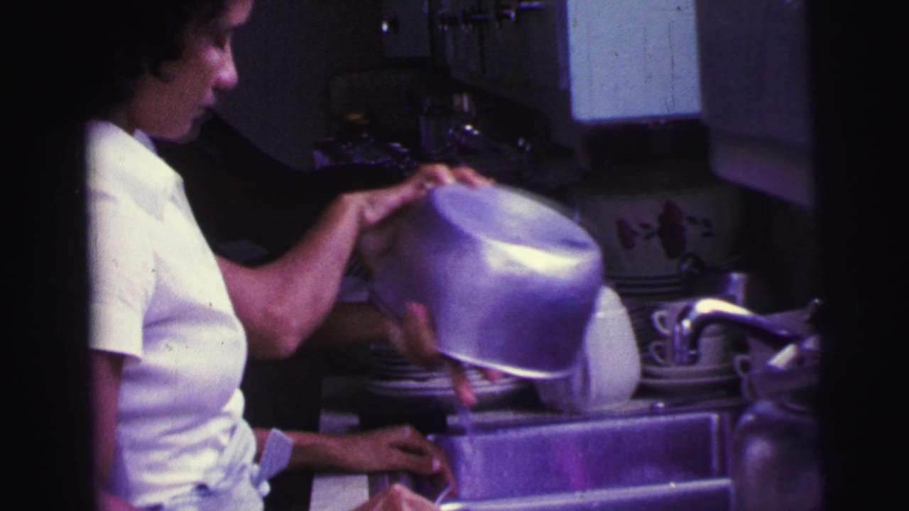 1967 Brazil 8mm frame of the woman holding a metal bowl over a crowded sink with stacked plates, cups, and a white teapot.