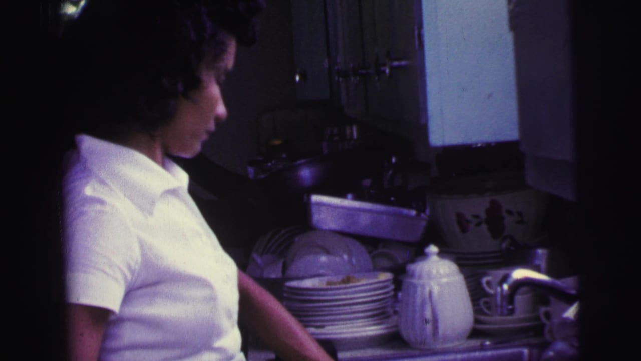 1967 Brazil 8mm frame showing the woman beside a dim kitchen sink with piled plates, cups, and a white teapot on the counter.