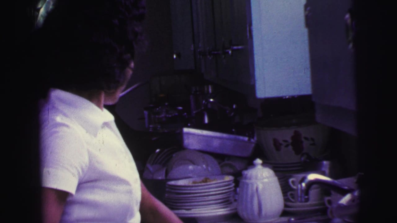Woman in a white collared shirt standing by a kitchen sink with stacked dishes.