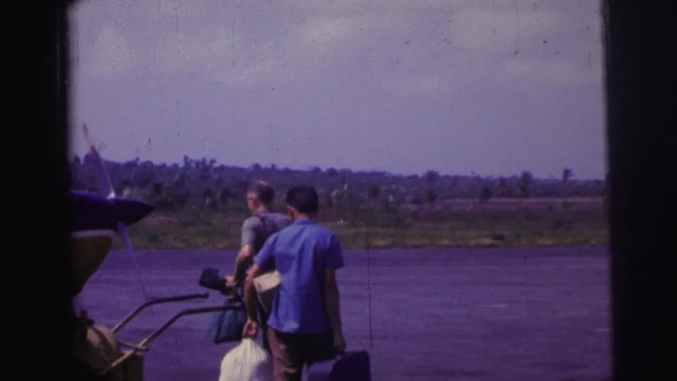 In 1967 Brazil, two men walk across a tarmac with luggage beside a parked small propeller plane.