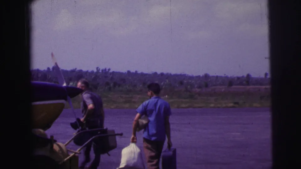 Men carrying luggage and gear cross an airfield in Brazil, with a small propeller plane parked nearby.