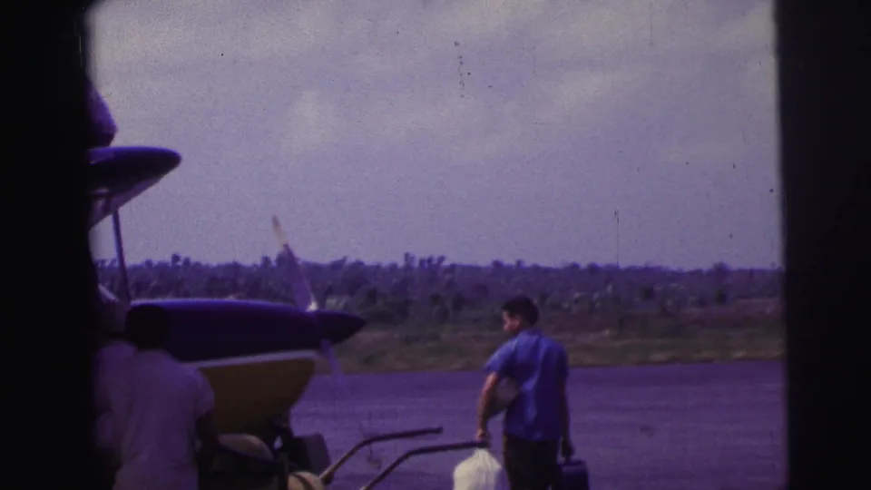A man in a blue shirt carries luggage across a Brazilian airfield as the aircraft nose fills the left side of frame.