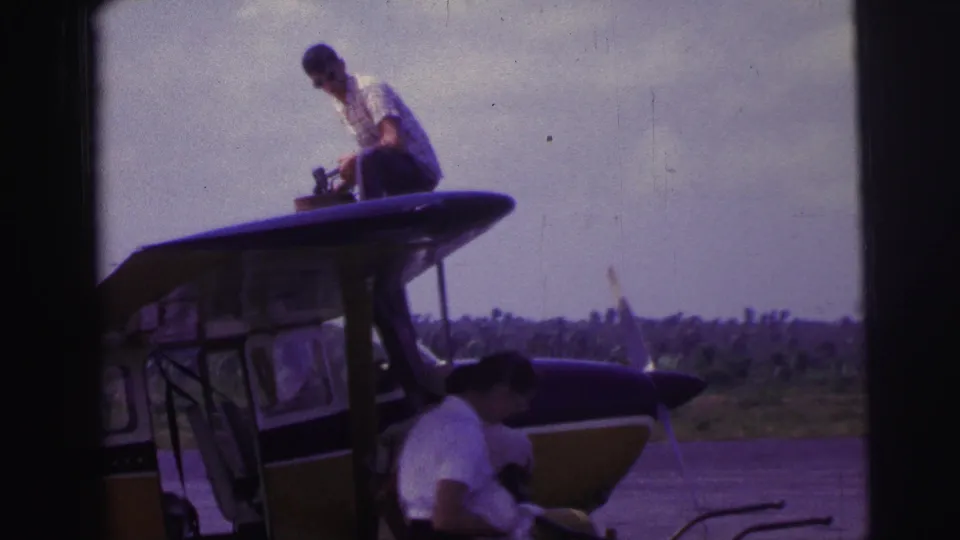 1967 Brazil 8mm frame showing two men around a small aircraft, one above the cabin and one below near the fuselage.