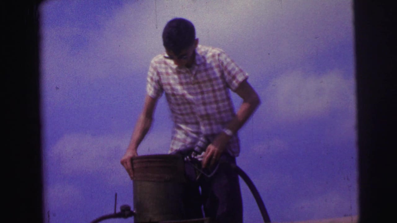 1967 Brazil home-movie frame of a man in sunglasses and a checkered shirt handling a bucket and equipment near the aircraft.