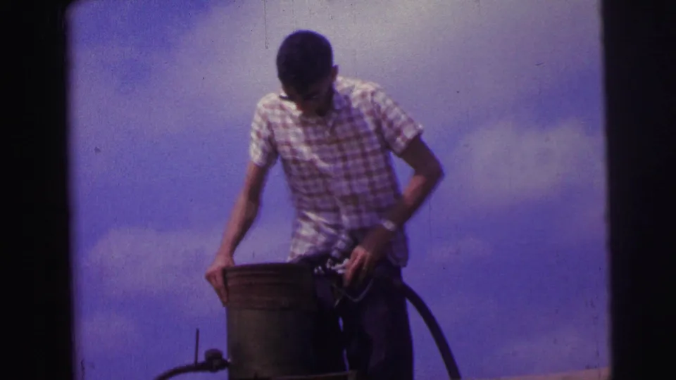 1967 Brazil home-movie frame of a man in sunglasses and a checkered shirt handling a bucket and equipment near the aircraft.
