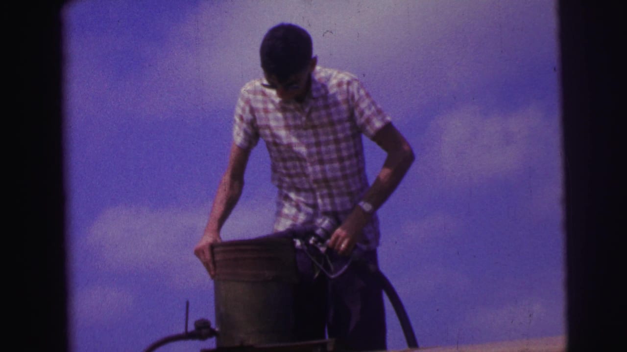 Man in checkered shirt holds a metal can beside a fuel hose under a blue sky.