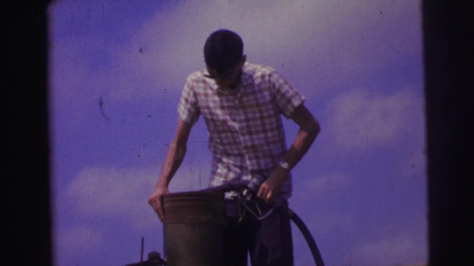 1967 Brazil home-movie frame showing a man looking down as he handles a fuel can and hose outdoors.