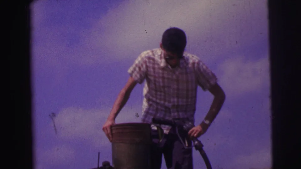1967 Brazil 8mm still of a man in a checkered shirt standing over a metal can while handling fuel equipment.